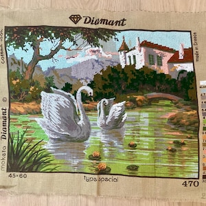 May include: A Diamant needlepoint canvas featuring a serene lake scene with two white swans, a building, and a bridge. The canvas measures 45 x 60 cm and is made in Greece. The text "Cotton 100%" is visible.