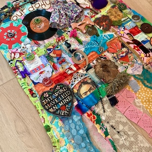 May include: A multi-textured fabric wall hanging featuring a collage of mixed media elements including a vinyl record, a floral dress, lace ribbons, a heart-shaped plaque with the text 'THY WORD HAVE I HID IN MINE HEART', and a portrait of Frida Kahlo.