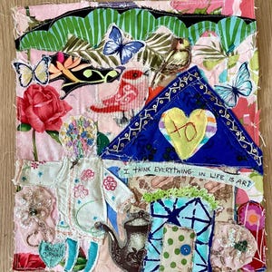 May include: A vibrant textile artwork with a patchwork design. The piece depicts a house shape with a blue roof and a yellow heart. Butterflies, birds, and floral patterns are included, along with the text "I think everything in life is art."