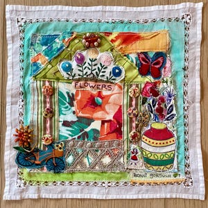 May include: A colorful textile art piece featuring a house with the word "FLOWERS" above a floral design. A butterfly, flowers, and a bicycle are also depicted. The artwork is framed with a white lace border and signed by "BONNI GORSUCH".