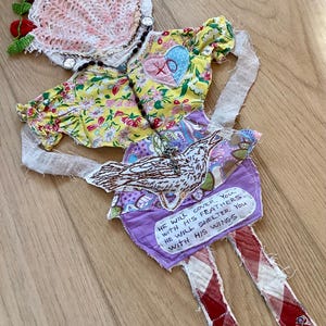 Puede incluir: Muñeca de tela hecha a mano con un vestido amarillo floral, un sombrero de ganchillo rosa y blanco con detalles rojos y piernas a cuadros rojos y blancos. La muñeca tiene una falda morada con un aplique de pájaro y una cita: "Él te cubrirá con sus plumas."