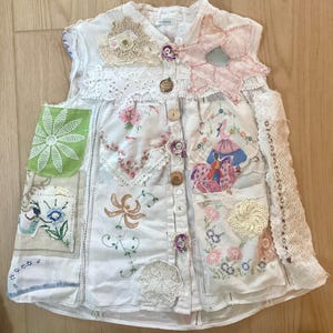 May include: A white patchwork vest with various fabric patches, lace, and embroidery. The vest features floral and geometric designs, with button closures and decorative buttons. The vest has a unique, handcrafted appearance.