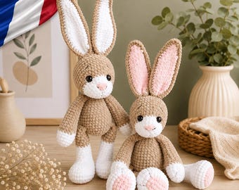 May include: Two crocheted rabbit dolls with tan bodies and white accents. The standing rabbit has long white-lined ears, while the sitting rabbit features pink-lined ears and pink paw pads. Both have black safety eyes and small pink noses.