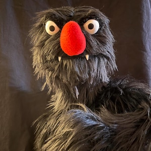 May include: A handmade puppet with a shaggy, dark gray fur body, large white eyes with black pupils, a bright red nose, and two small white fangs. The puppet has a friendly, monster-like appearance.