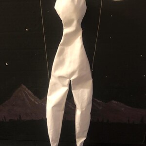 May include: A white puppet with a head, torso, and long legs, suspended by strings. The puppet is against a backdrop of a night scene with a moon, stars, and mountains.
