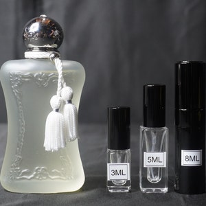 May include: Four perfume bottles, one large and three small. The large bottle is clear glass with a silver top and white tassels. The small bottles are clear glass with black caps and labels that say "3ML", "5ML", and "8ML".