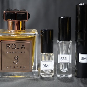 May include: Three perfume bottles with black caps and labels. The largest bottle is a clear glass bottle with a gold label that reads "ROJA PARFUMS 3 PARFUM". The smaller bottles are labeled "3ML", "5ML", and "8ML".