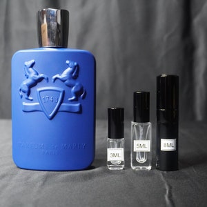 May include: A blue glass bottle of perfume with a silver cap and a crest featuring two horses. The bottle is labeled "Parfums de Marly" and "1743". Three smaller bottles of perfume are also shown, labeled "3ML", "5ML", and "8ML".