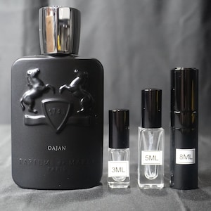 May include: A black bottle of Oajan perfume with a silver cap. The bottle is decorated with two horses and a shield with the number 174. The bottle is next to three smaller bottles of perfume, one 3 ml, one 5 ml, and one 8 ml.