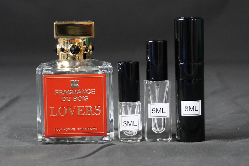 Op de afbeelding: Een heldere glazen parfumfles met een gouden dop en zwarte accenten, met het opschrift "FRAGRANCE DU BOIS LOVERS" in gouden letters op een rode achtergrond. Er zijn ook drie kleinere parfumflesjes afgebeeld met 3 ml, 5 ml en 8 ml.