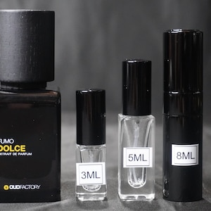 May include: Four bottles of perfume in different sizes. The largest bottle is black with a wooden cap and the label "Fumo Dolce Extrait de Parfum" and the logo "OUDFACTORY". The other three bottles are smaller and have black caps. The bottles are labeled with the volume in milliliters: 3ML, 5ML, and 8ML.