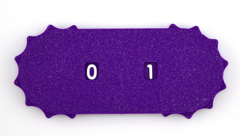 May include: A purple, textured counter with a unique, starburst-like edge. The counter displays the numbers "0" and "1" in white, centered within rectangular cutouts. The object is likely used for counting or tracking.