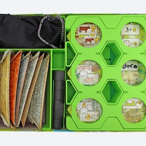 Puede incluir: Una caja de juego de mesa abierta con un interior verde, que contiene piezas de juego, cartas y una bolsa negra con cordón. Las cartas tienen varios colores y texturas. Las piezas del juego incluyen cilindros negros y fichas circulares.
