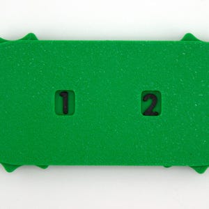 May include: A bright green, rectangular foam object with a textured surface. The object has a series of rounded edges and two cut-out squares with the numbers "1" and "2" in black. The object is on a white background.