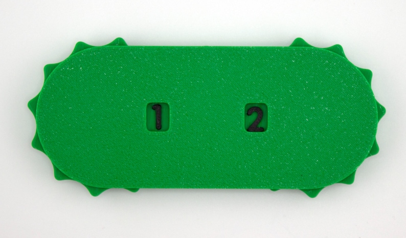 May include: A bright green, rectangular foam object with a textured surface. The object has a series of rounded edges and two cut-out squares with the numbers "1" and "2" in black. The object is on a white background.