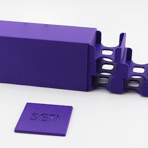 May include: A purple card holder with a matching box and a small square with the word "SETI" embossed on it. The card holder has a stepped design with multiple slots for holding cards.