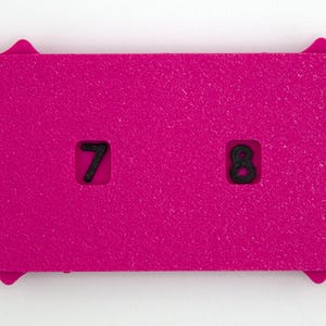May include: A bright pink, rectangular object with a textured surface. The object has a decorative, sunburst-like edge. Two black numbers, 7 and 8, are cut out of the pink surface. The object is likely a game piece or a counter.