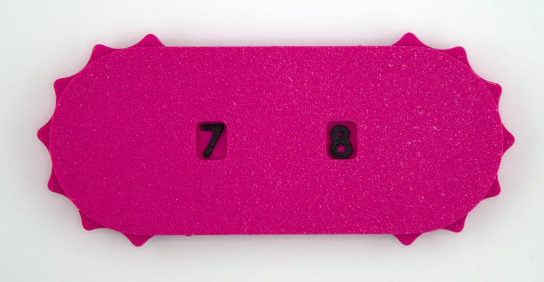 May include: A bright pink, rectangular object with a textured surface. The object has a decorative, sunburst-like edge. Two black numbers, 7 and 8, are cut out of the pink surface. The object is likely a game piece or a counter.