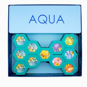 May include: AQUA board game in a blue box. The box lid displays the word "AQUA" in blue. Inside, teal hexagonal tiles with colorful ocean-themed images are arranged. The game appears to be a puzzle or strategy game.