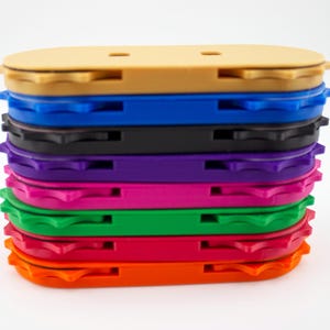May include: A stack of colorful, oval-shaped plastic objects. The objects are in various colors, including tan, blue, black, purple, pink, green, and orange. Each object has a grooved edge and a small slot on the top.