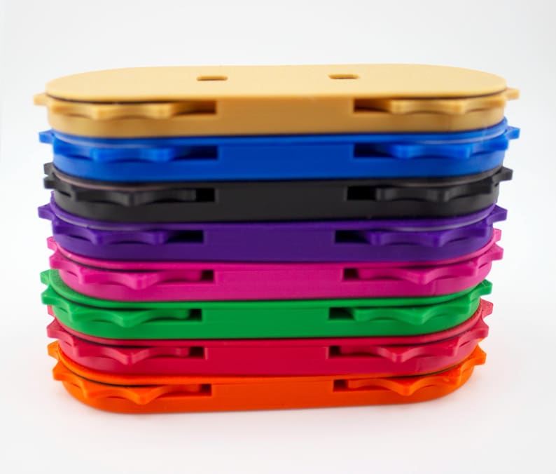 May include: A stack of colorful, oval-shaped plastic objects. The objects are in various colors, including tan, blue, black, purple, pink, green, and orange. Each object has a grooved edge and a small slot on the top.