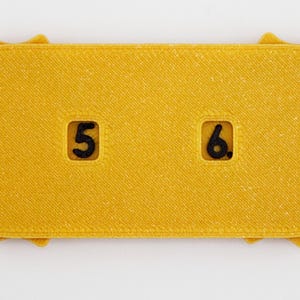 May include: A yellow, rectangular object with rounded ends and a textured surface. The object has a sun-like edge design. It features the numbers "5" and "6" in black, within recessed squares. The item appears to be a game piece or a decorative element.