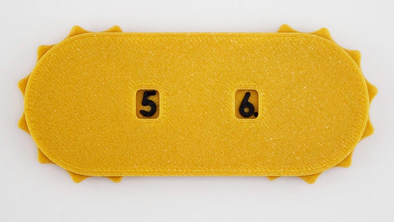 May include: A yellow, rectangular object with rounded ends and a textured surface. The object has a sun-like edge design. It features the numbers "5" and "6" in black, within recessed squares. The item appears to be a game piece or a decorative element.