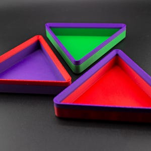 Puede incluir: Tres bandejas triangulares coloridas. Cada bandeja tiene un borde y un interior contrastantes, creando un diseño geométrico vibrante. Las bandejas son rojas, moradas y verdes, dispuestas sobre una superficie oscura.