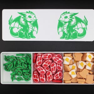 May include: A white rectangular box with a green dragon design and a three-compartment tray filled with green leaf shapes, red meat shapes, and toast shapes with butter. The dragon design is repeated twice.