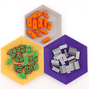 May include: Three hexagonal trays in yellow, grey, and purple, each filled with game pieces. The yellow tray holds green leaf shapes, the grey tray has orange barrel shapes with blue lightning bolts, and the purple tray contains grey rectangular pieces.