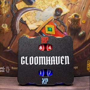 Könnte beinhalten: Ein schwarzer Gloomhaven-Spiel-Tracker aus Kunststoff mit strukturierter Oberfläche. Das Gerät verfügt über zwei Reihen kreisförmiger Ausschnitte, die rote und blaue nummerierte Zifferblätter zeigen. HP ist in Rot darüber und XP in Blau darunter gedruckt.