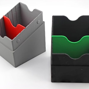 Könnte beinhalten: Zwei Kartenaufbewahrungsboxen. Eine ist grau mit einem roten Teiler, die andere ist schwarz mit einem grünen Teiler. Beide haben ein schräges Design und eine gebogene obere Öffnung. Die Boxen sind für Karten konzipiert.