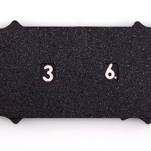 May include: A black, textured, rectangular object with a scalloped edge. The numbers "3" and "6" are displayed in a light pink color. The object is set against a white background.
