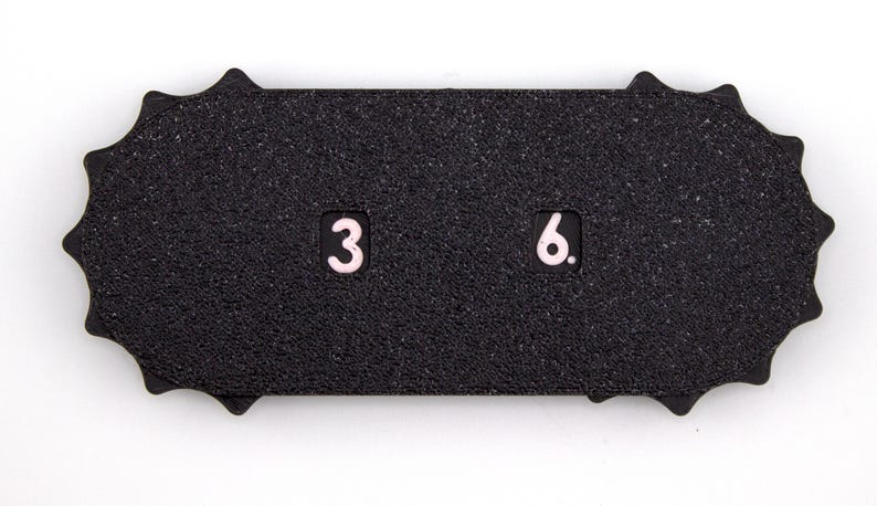 May include: A black, textured, rectangular object with a scalloped edge. The numbers "3" and "6" are displayed in a light pink color. The object is set against a white background.