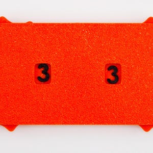 May include: Bright orange rectangular object with rounded ends and a textured surface. Two black numbers "3" are visible. The ends have a sunburst design. The object is likely a game piece or a counter.