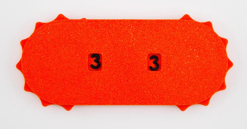 May include: Bright orange rectangular object with rounded ends and a textured surface. Two black numbers "3" are visible. The ends have a sunburst design. The object is likely a game piece or a counter.