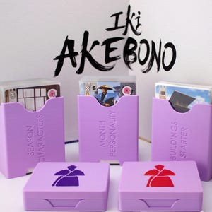 Könnte beinhalten: Eine Sammlung von lilafarbenen Spielkartenhaltern und -boxen. Die Halter sind mit "Season Characters", "Month Personality" und "Buildings Starter" beschriftet. Die Boxen zeigen ein Sumo-Ringer-Design. Der Text "Iki Akebono" steht in Schwarz darüber.