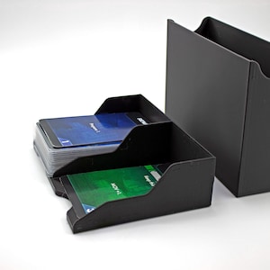 Puede incluir: Juego de cajas negras para guardar cartas. El juego incluye una caja rectangular alta y dos tarjeteros de dos niveles. Los tarjeteros están llenos de cartas con diseños azules y verdes. Las cajas son de material negro mate.