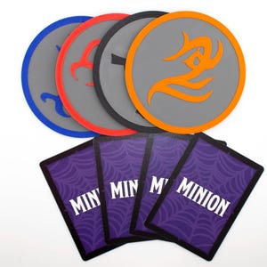 May include: Four circular coasters with grey centres and coloured borders in blue, red, black, and orange, each featuring a unique symbol. Below them are four rectangular playing cards with purple spiderweb patterns and the word MINION printed.