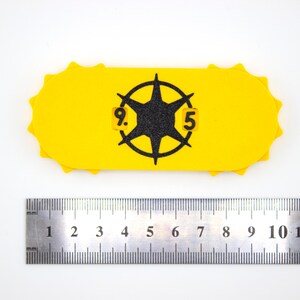 May include: A yellow, oblong-shaped object with a black starburst design and the numbers "9.5" in a circle. The object has a scalloped edge. A metal ruler is placed below the object, showing measurements in inches.
