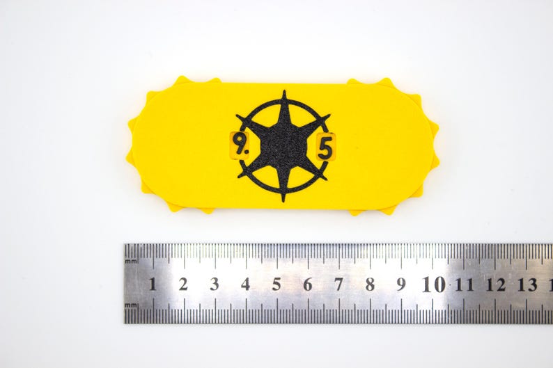 May include: A yellow, oblong-shaped object with a black starburst design and the numbers "9.5" in a circle. The object has a scalloped edge. A metal ruler is placed below the object, showing measurements in inches.
