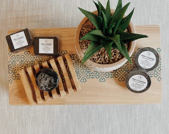 May include: A wooden tray with a green and yellow patterned border. There are three black charcoal soap bars, one in a bamboo soap dish with a white lather, and a potted succulent plant in the center.
