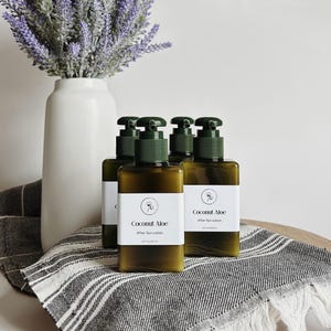 May include: Four olive-green bottles of Coconut Aloe After Sun Lotion with dark green pump dispensers are arranged on a striped towel. A white vase with lavender stems is in the background. The bottles each contain 3.3 fl oz / 100 mL.