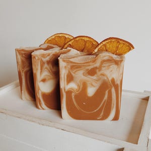 May include: Three rectangular bars of soap featuring a marbled pattern of cream and burnt orange colors. Each bar is topped with a single, dehydrated orange slice embedded into the top edge of the soap, creating a textured finish.