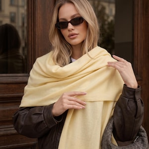 May include: A soft, pale yellow rectangular scarf draped over the shoulders, paired with a dark brown leather jacket, a woven dark grey hobo bag, and dark-tinted oval sunglasses. The scarf features a smooth, solid-coloured knit texture.