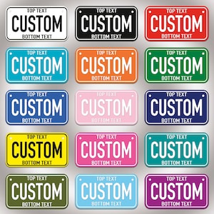 Peut inclure: Une collection de quinze plaques d'immatriculation rectangulaires de différentes couleurs unies, notamment blanc, noir, rouge, bleu, orange, vert, jaune, rose et violet. Chaque plaque porte le texte TOP TEXT, CUSTOM et BOTTOM TEXT en gras.