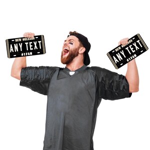 Puede incluir: Una persona con una camiseta negra sostiene dos carteles negros y dorados con forma de matr&iacute;cula. Los carteles dicen "NEW ORLEANS ANY TEXT #1 FAN".