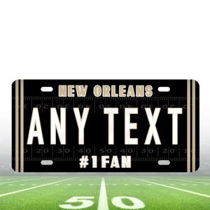 Puede incluir: Una placa de matr&iacute;cula personalizada negra y dorada con el texto "NEW ORLEANS" y "ANY TEXT" en blanco. La parte inferior de la placa dice "#1 FAN".