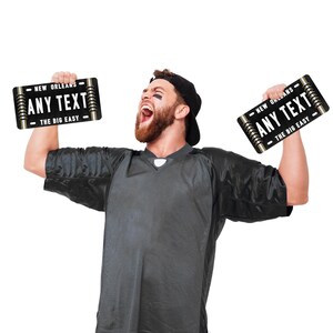 Puede incluir: Una persona con una camiseta negra sostiene dos carteles negros y dorados con el texto "NEW ORLEANS ANY TEXT THE BIG EASY" en ellos.
