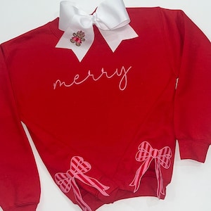 Pode incluir: Camisola vermelha com a palavra "merry" bordada em branco. A camisola tem laços listrados rosa e branco nas mangas e um laço branco com um pequeno aplique de urso de peluche no pescoço.